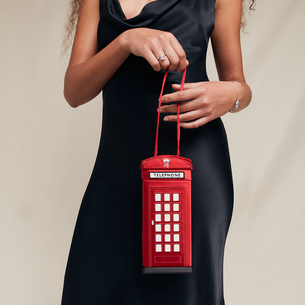 Red phone booth-shaped handbag held by a person wearing a black dress.