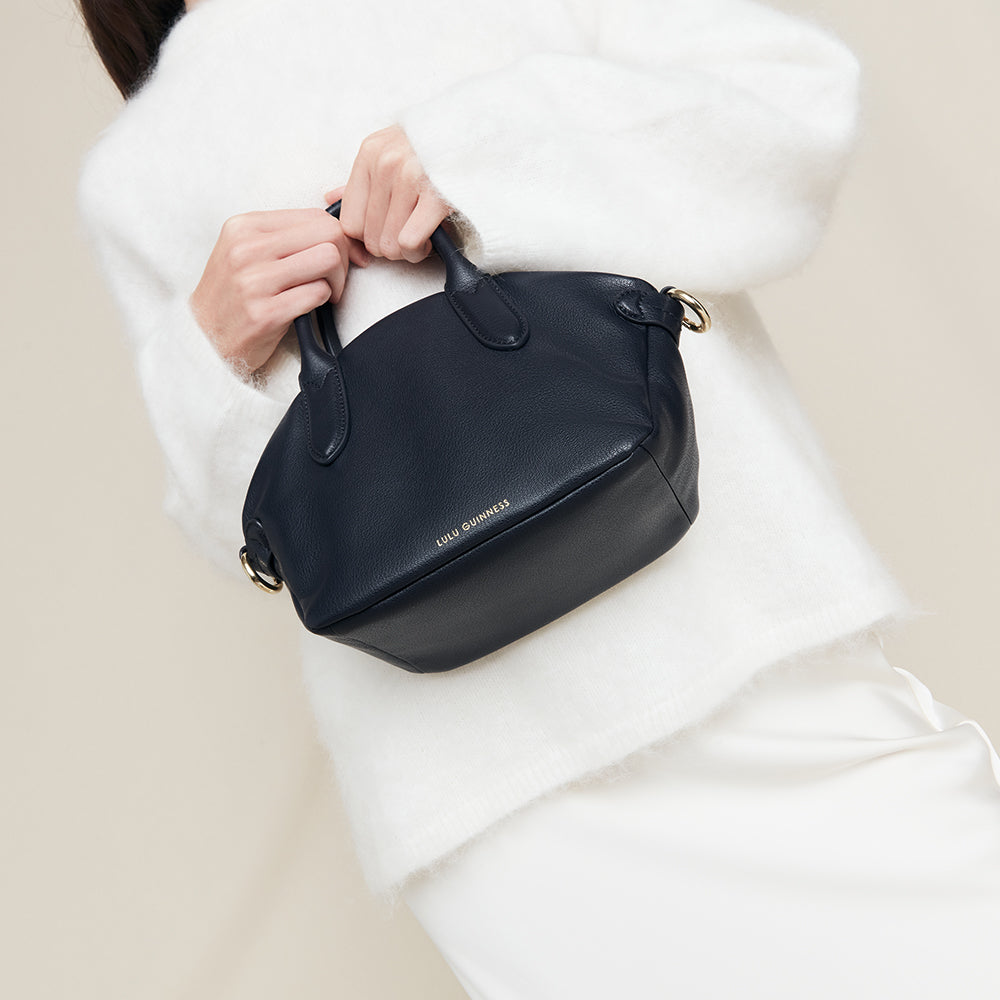 Person holding a small navy leather  handbag with 'Lulu Guinness' branding against a neutral background