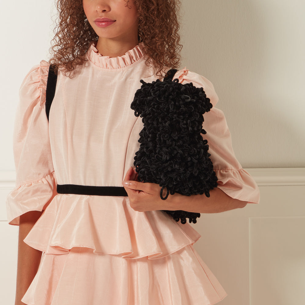 A woman with curly brown hair wears a light pink tiered dress and holds a black fluffy handbag.