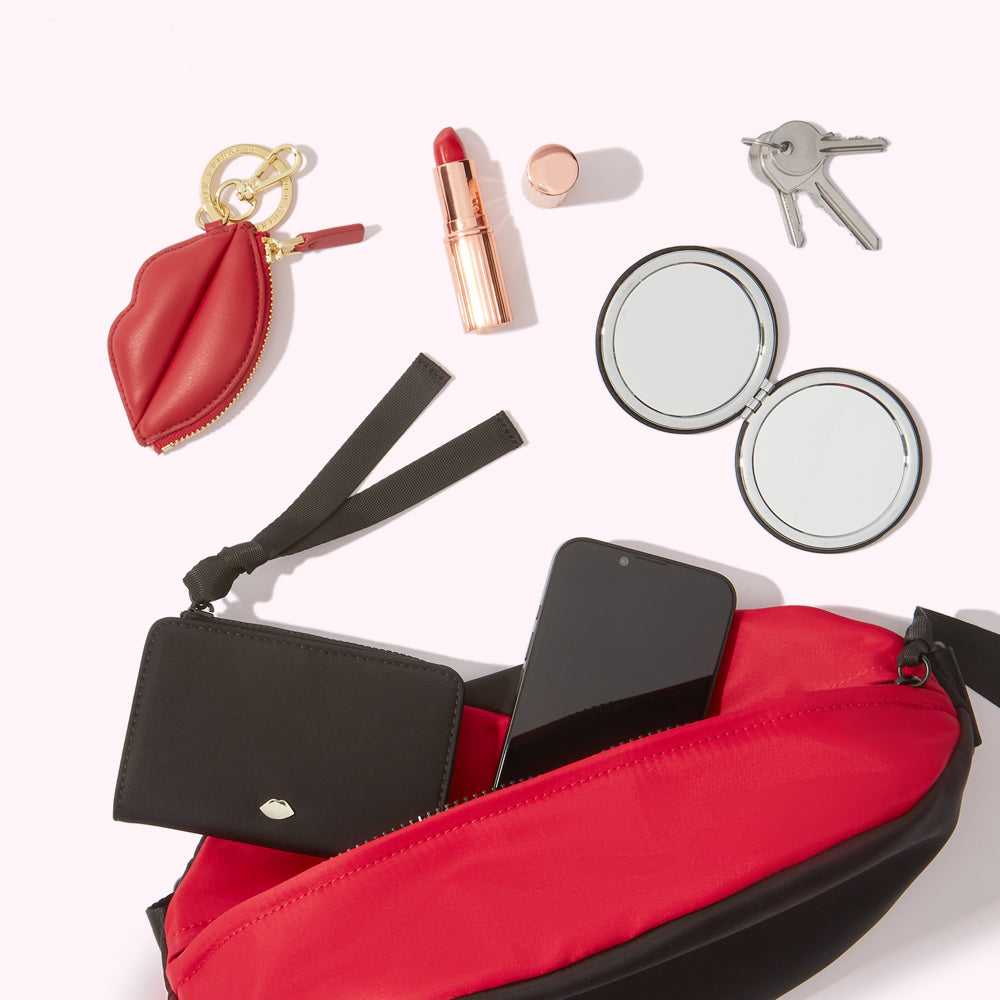 Red and black belt bag with black interior containing a black wallet, smartphone, and a compact mirror; also shown are a red lip-shaped keychain, lipstick, and keys.
