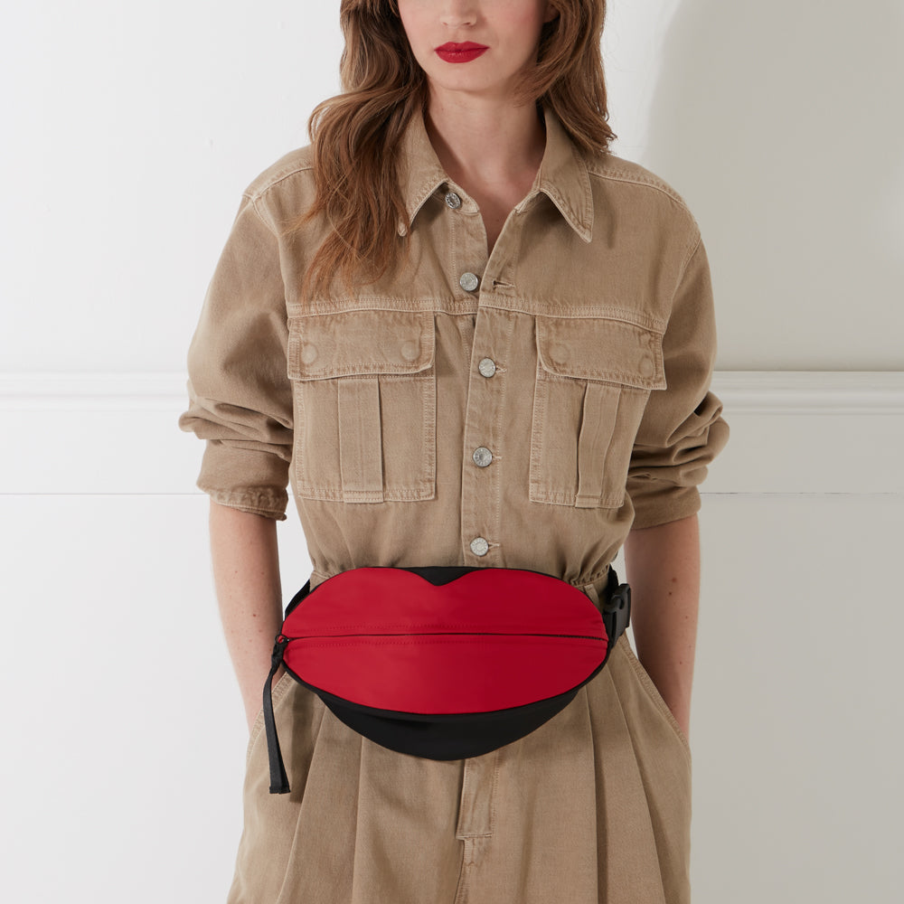 A woman in a tan denim jumpsuit wears a red lip-shaped belt bag.
