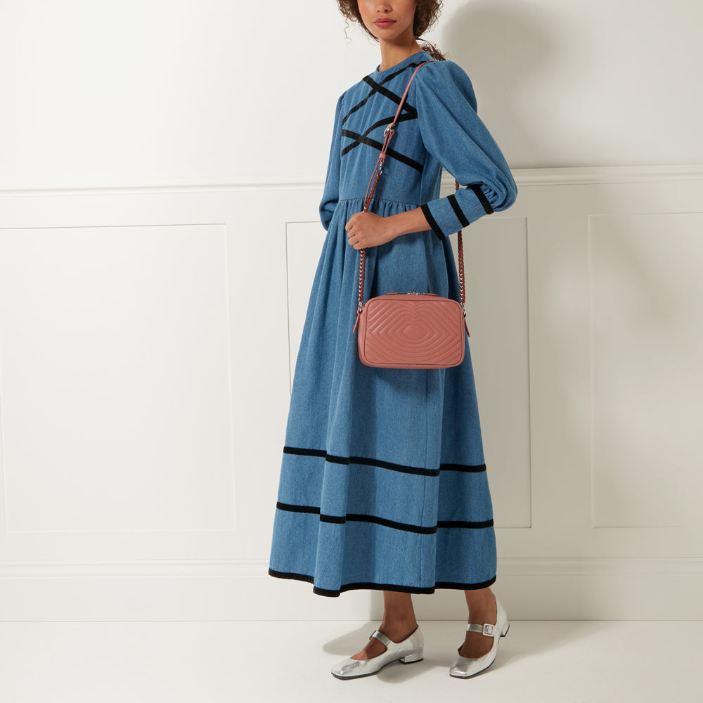A woman in a denim dress carries a dusty rose quilted crossbody bag with a silver chain strap against a white background.
