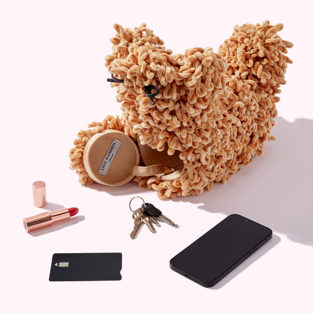 A fluffy, light brown Lulu Guinness dog-shaped bag sits on a white background next to a lipstick, keys, and a smartphone. The bag is partially open, revealing a small beige pouch inside. A black credit card is also visible.
