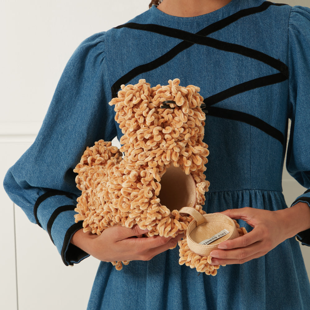 Person in a denim dress holding a tan fluffy dog-shaped purse. The purse is open to reveal a small, beige interior.
