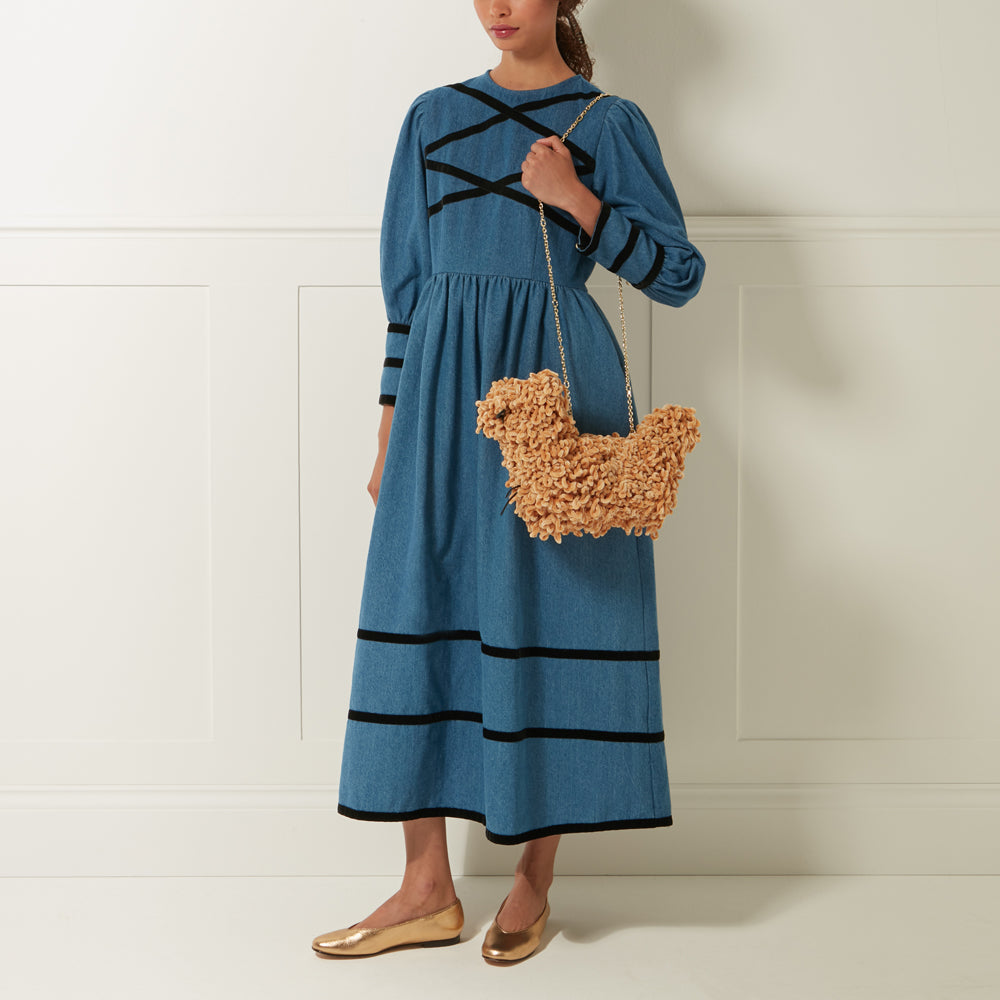A woman in a denim dress and gold flats carries a tan, fluffy dog-shaped purse with a gold chain strap.
