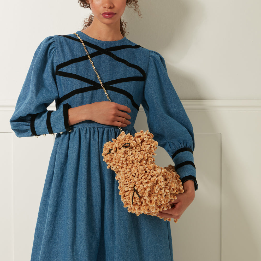 A woman in a denim dress holds a tan, fluffy teddy bear-shaped bag with a gold chain strap.