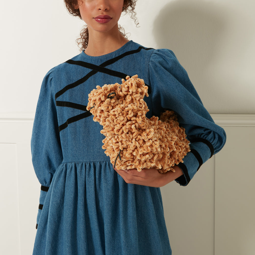 A person in a blue denim dress holds a tan, fluffy, dog-shaped bag.
