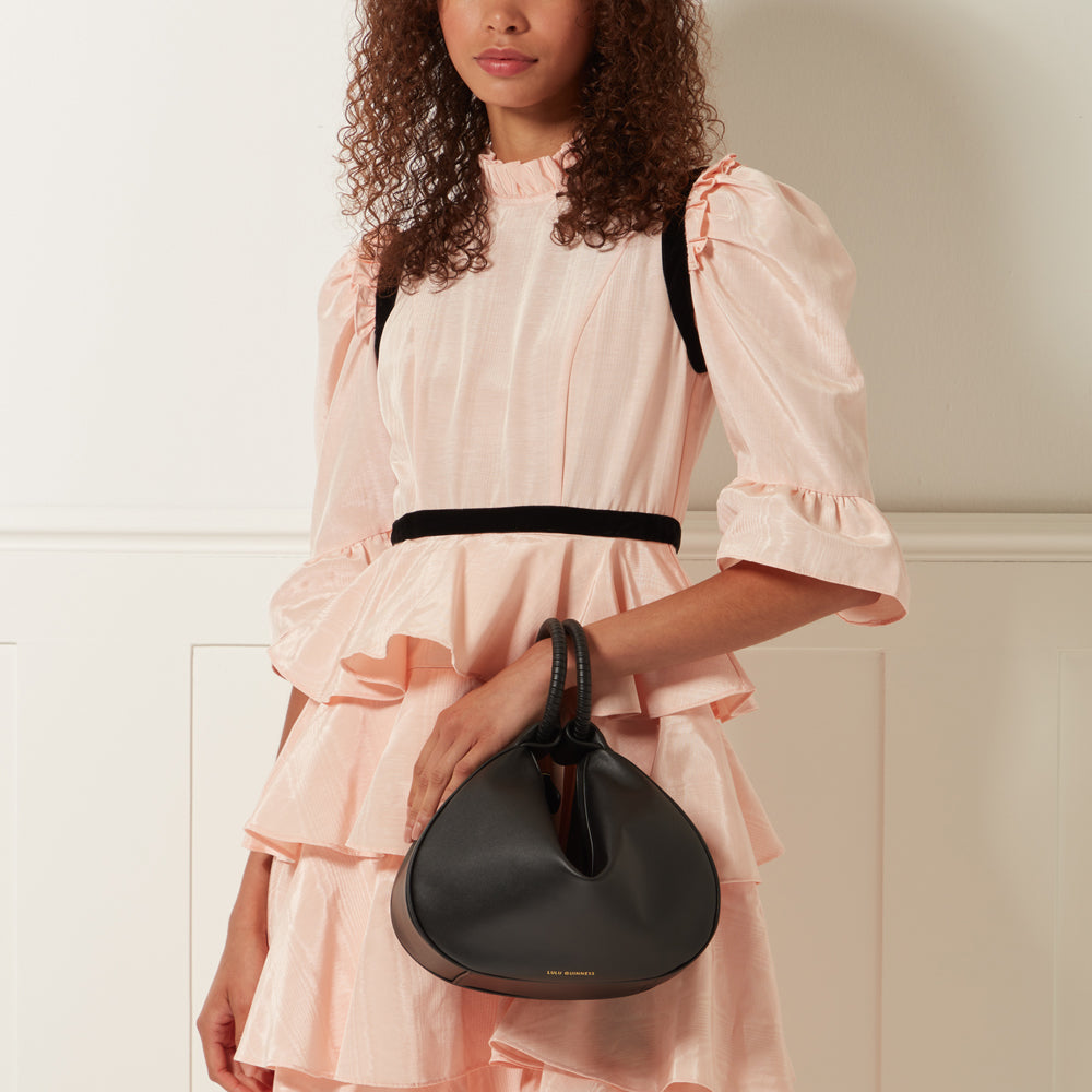A woman in a peach dress holds a black Lulu crossbody bag.
