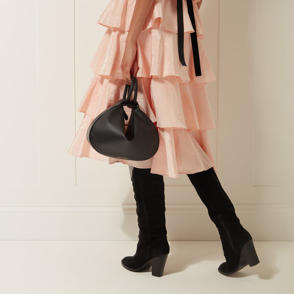 A woman in a pink dress and black boots carries a black Lulu Guinness bag.
