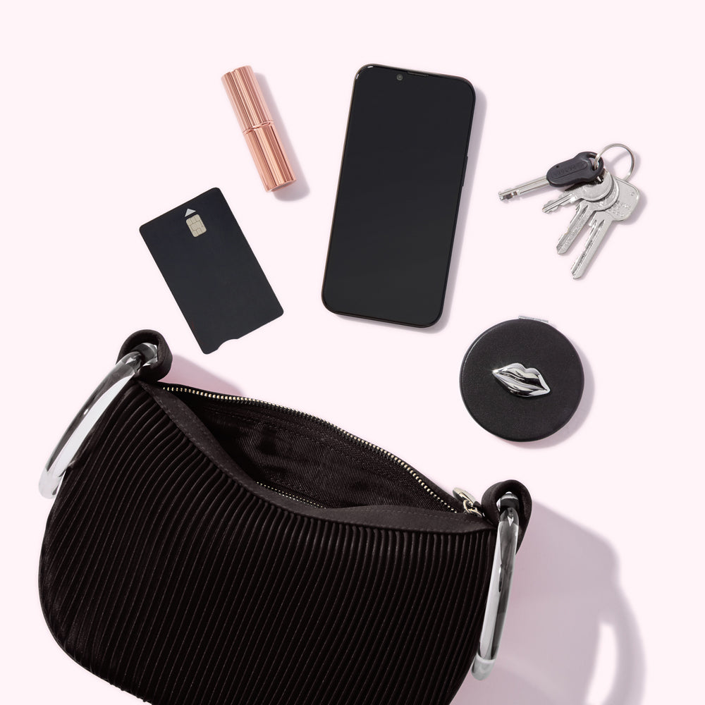 A black handbag sits open, displaying its pleated interior. Surrounding it are a smartphone, lipstick, credit card, keys, and a compact mirror with a lip design. The items are arranged on a light pink background.
