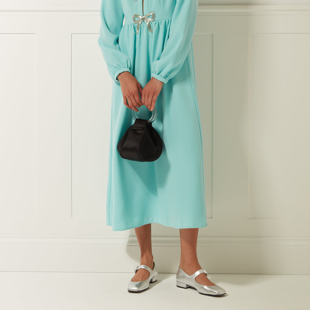 Woman in teal midi dress holding a black pleated handbag with a clear handle and wearing silver shoes against a white wall.
