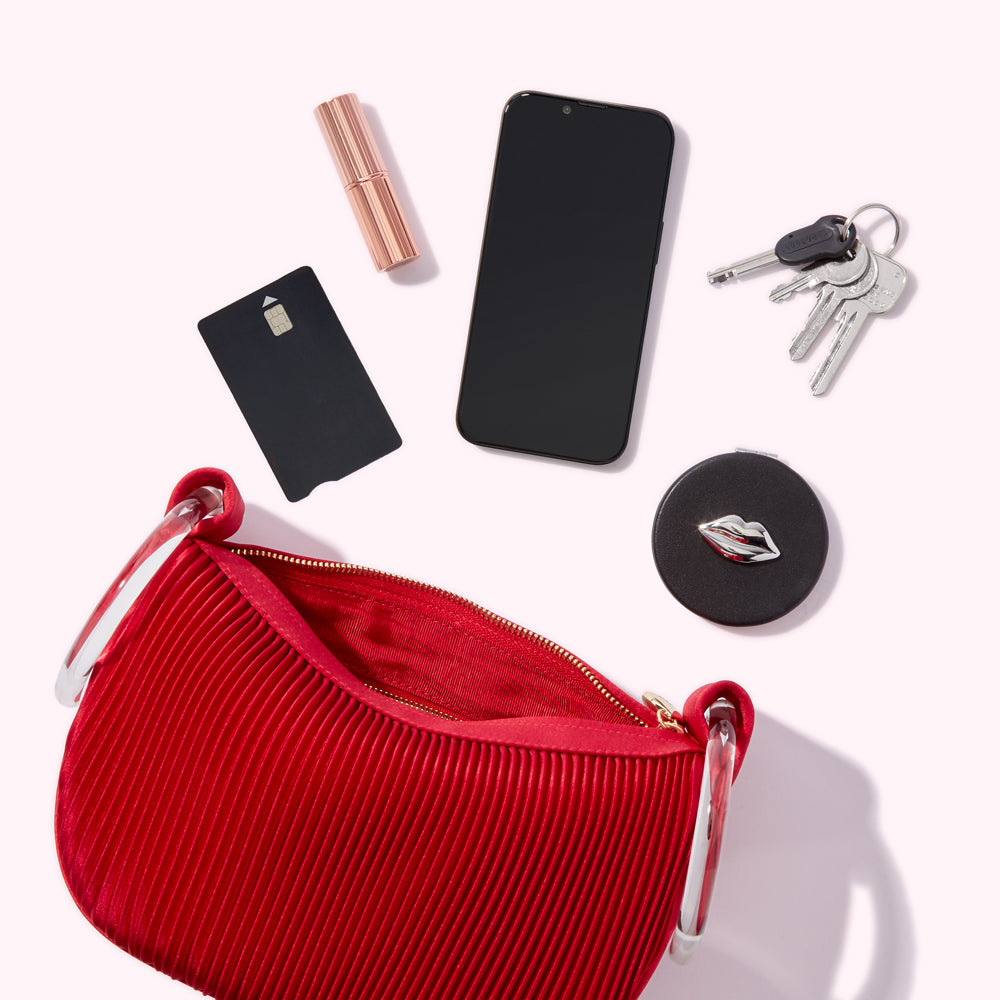 Red pleated handbag with a clear handle, next to a smartphone, lipstick, credit card, keys, and a compact mirror. The background is white.

