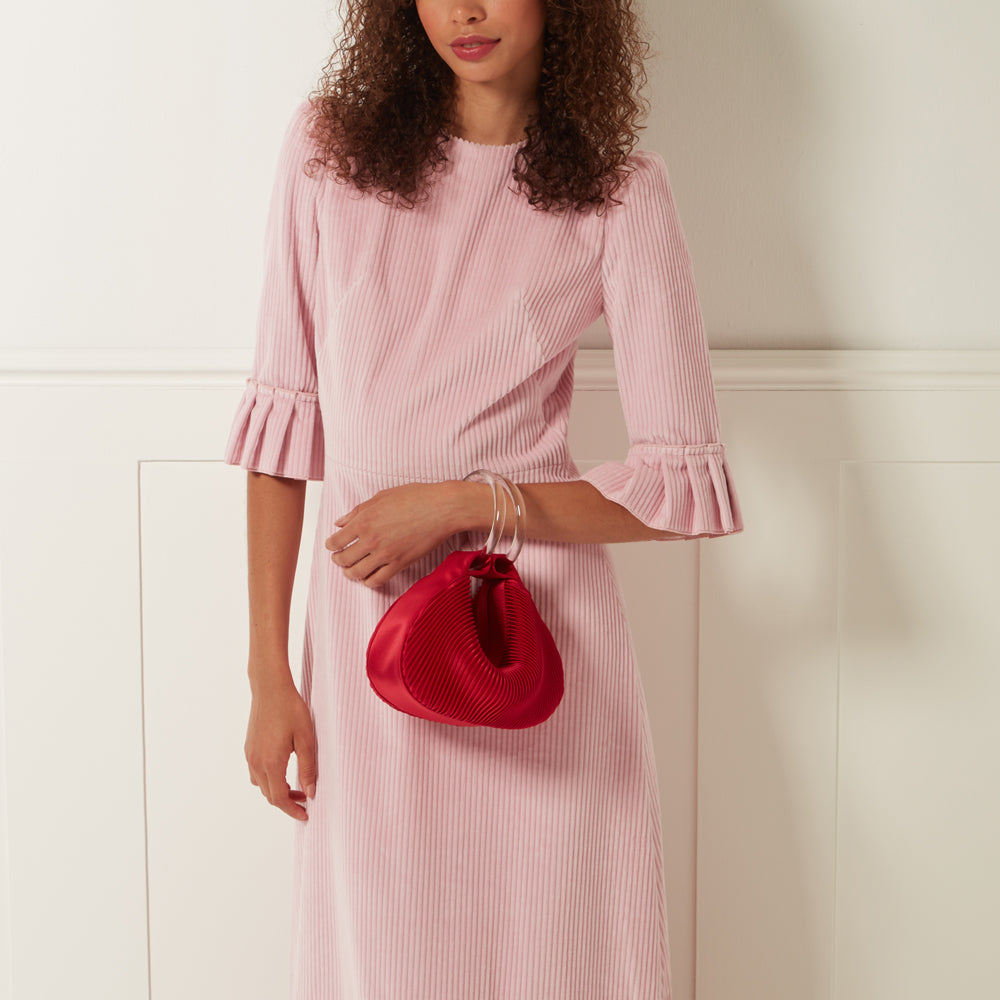 Woman in pink corduroy dress holding a red pleated satin bag with a clear handle against a white background.
