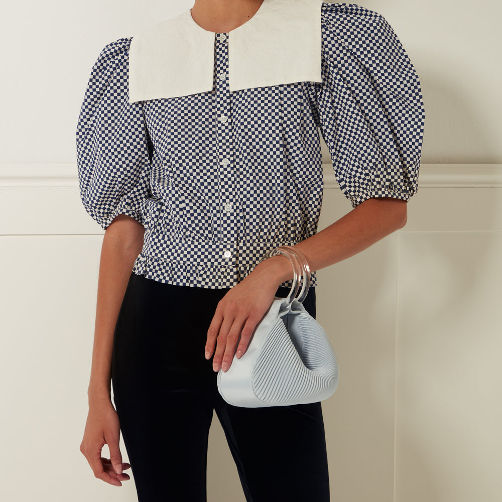 A woman with dark hair is modelling a blue and white gingham puff-sleeve blouse with a large white Peter Pan collar, black velvet pants, and a light blue pleated handbag with clear acrylic handles. The background is off-white.
