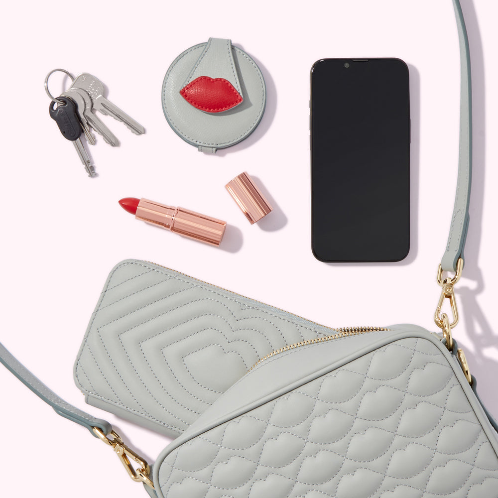 A light grey quilted handbag with a matching wallet, keys, a compact mirror with a red lip design, lipstick, and a smartphone.