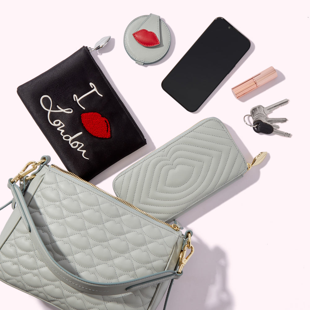 A light green quilted handbag, a black cosmetic bag that says "I love London", a light green quilted wallet, a compact mirror, a smartphone, a lipstick, and a set of keys are arranged on a white background.
