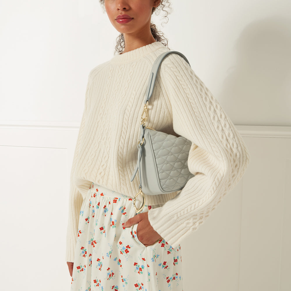 A woman wearing a cream-colored cable knit sweater and a floral skirt carries a light blue quilted shoulder bag.