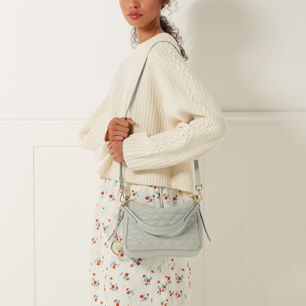 A woman in a cream-colored cable knit sweater and floral skirt carries a light blue quilted crossbody bag.

