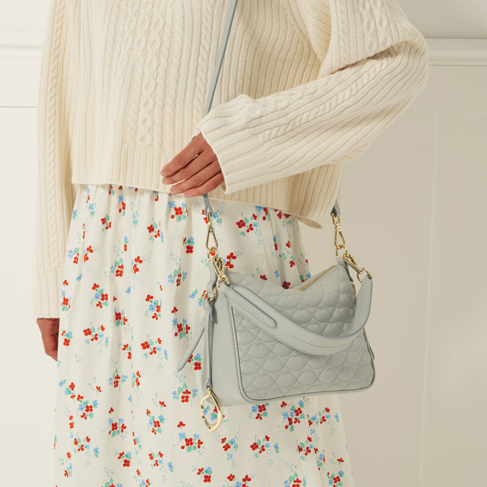 A light blue quilted crossbody bag with gold hardware is worn by a model. The model is wearing a cream-colored cable knit sweater and a floral print skirt.