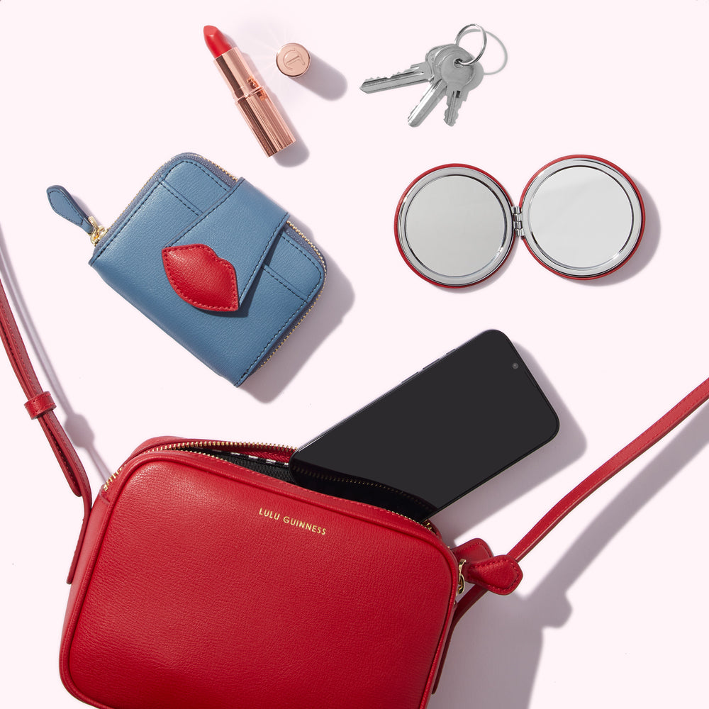 A red crossbody bag with a phone, keys, compact mirror, lipstick and a blue wallet on a white background.