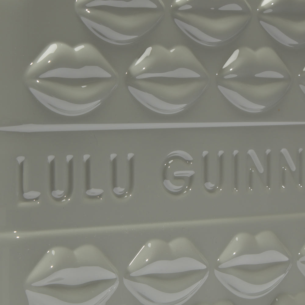 Close-up of a pale grey Lulu Guinness item featuring a repeating pattern of embossed lips and the brand name.