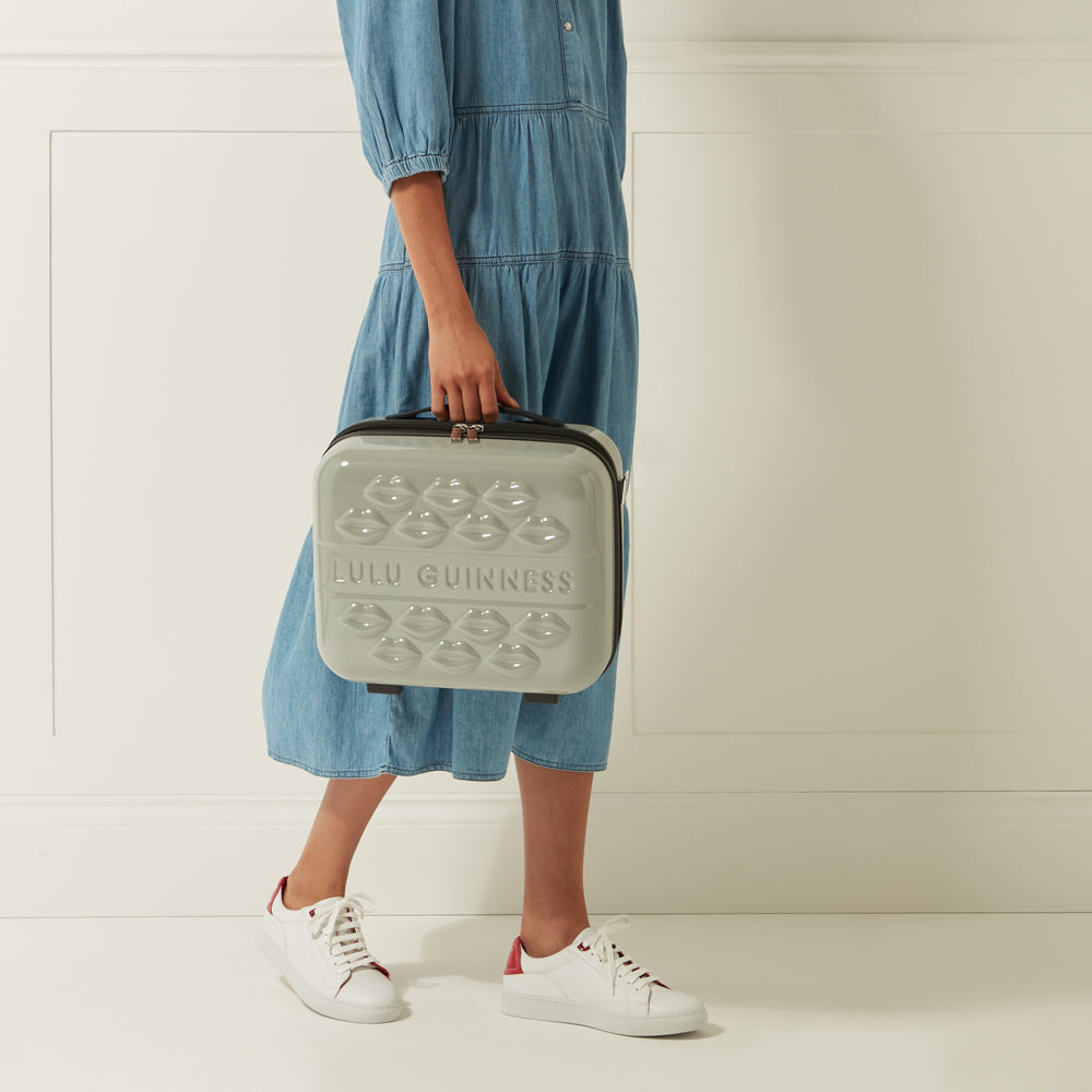 A woman in a denim dress holds a light grey Lulu Guinness makeup case decorated with embossed lips.
