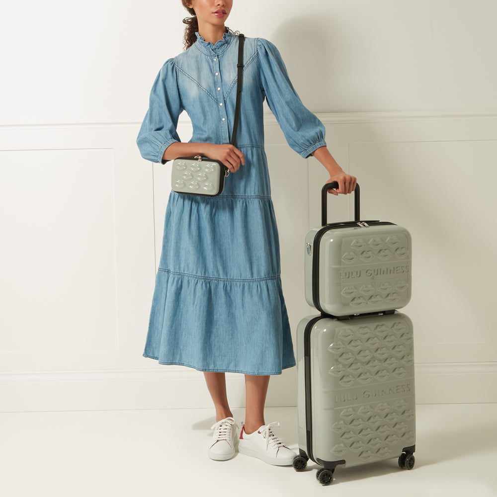 Woman in denim dress holding a Lulu Guinness kissing lips crossbody bag and matching luggage set against a white background.