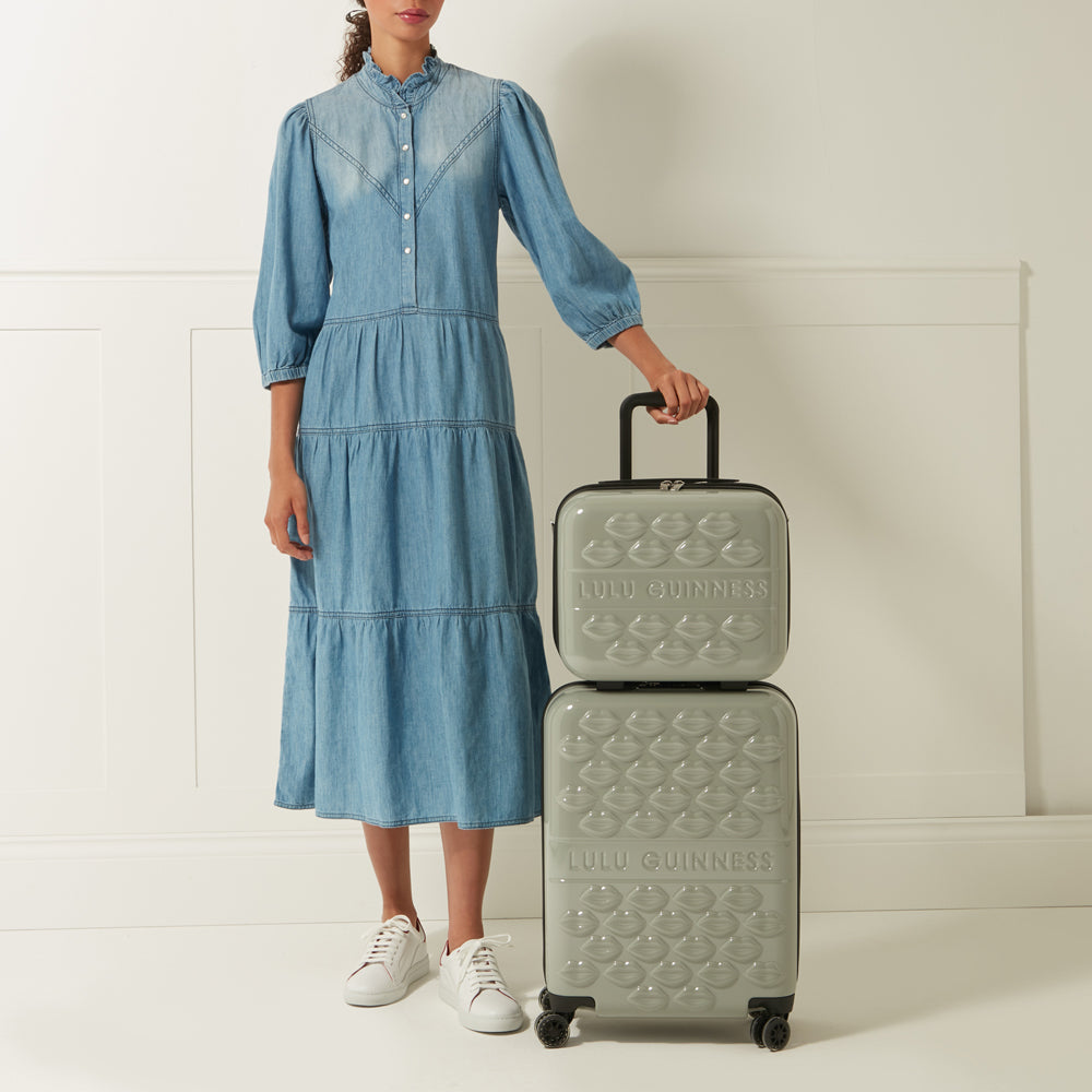 A woman in a denim dress stands next to a pale sage green Lulu Guinness two-piece luggage set decorated with a lip print.
