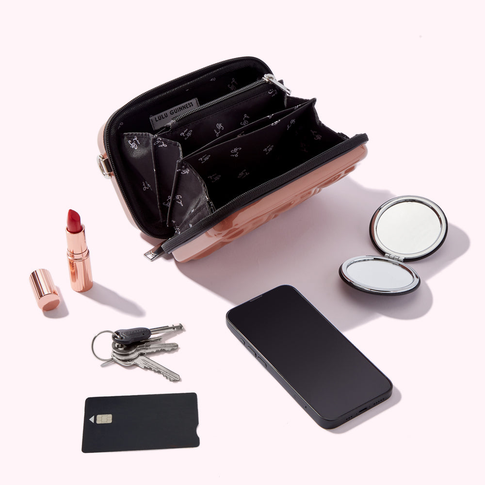 A rose gold Lulu Guinness purse is open, revealing its interior compartments. Beside it are a lipstick, a compact mirror, a smartphone, and a set of keys and a credit card. All items are on a white background.