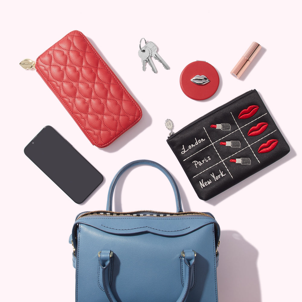 A blue handbag with a red quilted wallet, a black makeup bag with lipstick and city names embroidered on it, keys, a compact mirror, a phone, and a lipstick are arranged on a white background.