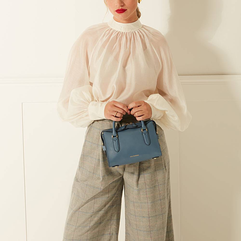 A woman in a white blouse and grey plaid pants holds a small blue leather handbag.
