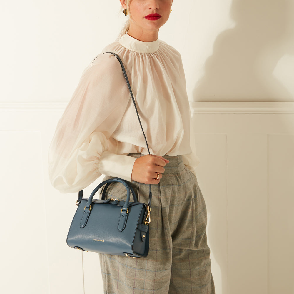 Woman in a white blouse and grey trousers holding a dark blue satchel handbag with gold detailing. The background is off-white.
