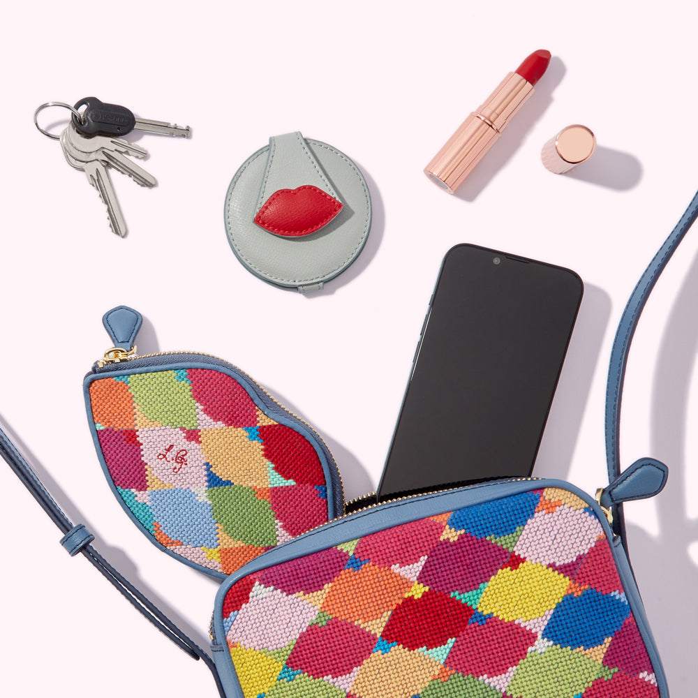 A colourful crossbody bag with a lip-shaped purse, keys, a compact mirror, lipstick, and a smartphone spilling out of it against a white background.