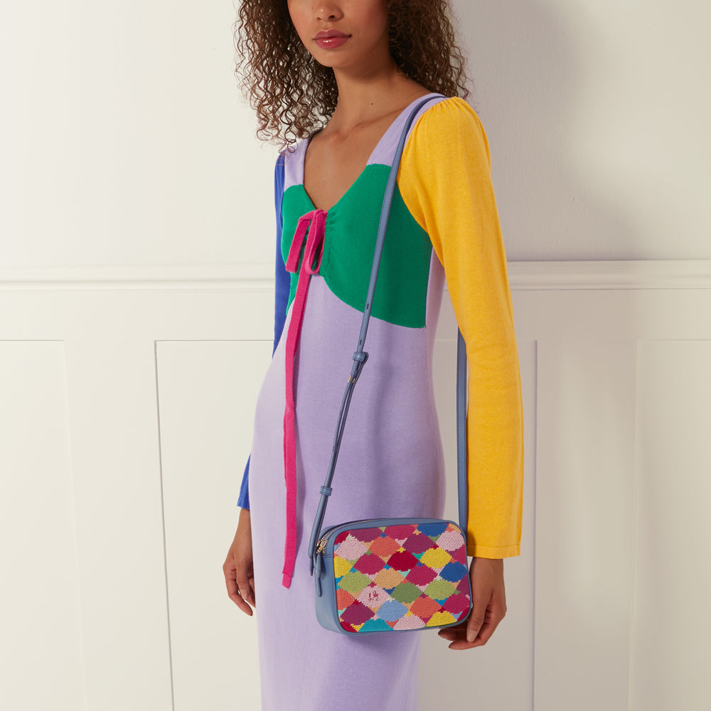 A woman in a colourful knit dress carries a small, light blue crossbody bag with a multi-coloured embroidered pattern against a white background.

