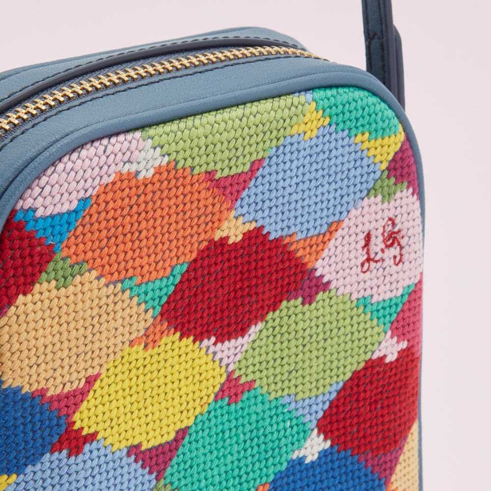 Close-up of a small, rectangular crossbody bag with a vibrant multi-coloured needlepoint pattern and a dark blue leather trim.