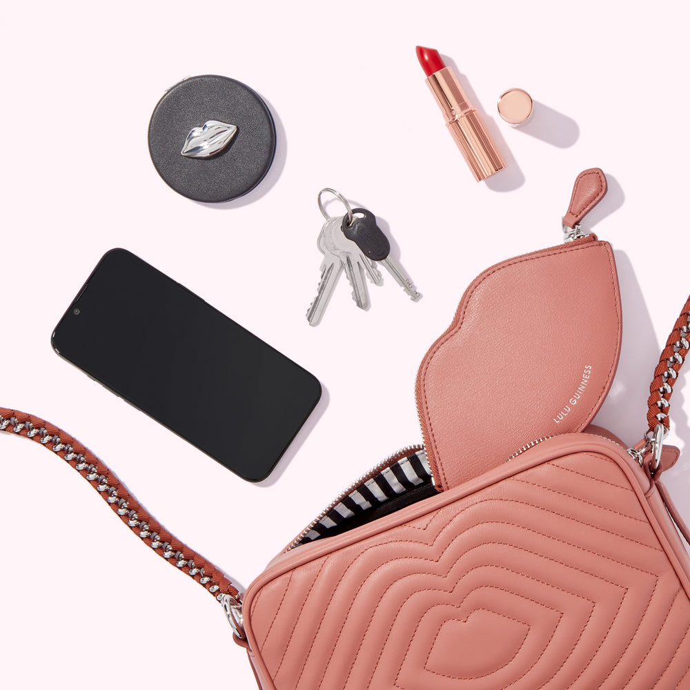 A blush pink quilted Lulu Guinness crossbody bag with a lip-shaped pouch inside, accompanied by a smartphone, keys, lipstick, and a compact mirror, all arranged on a white background.
