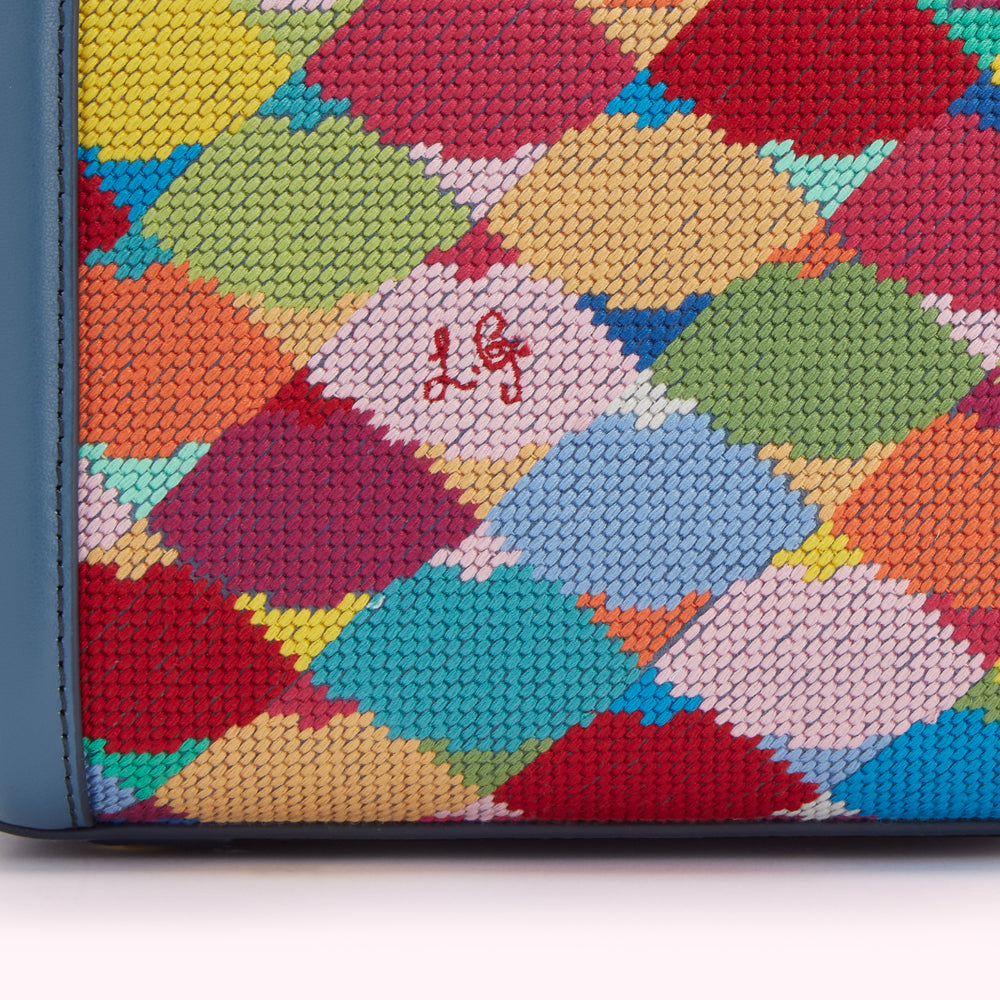 Close-up of a colourful needlepoint textile with a blue leather border, featuring a multi-coloured, abstract pattern and subtle logo.