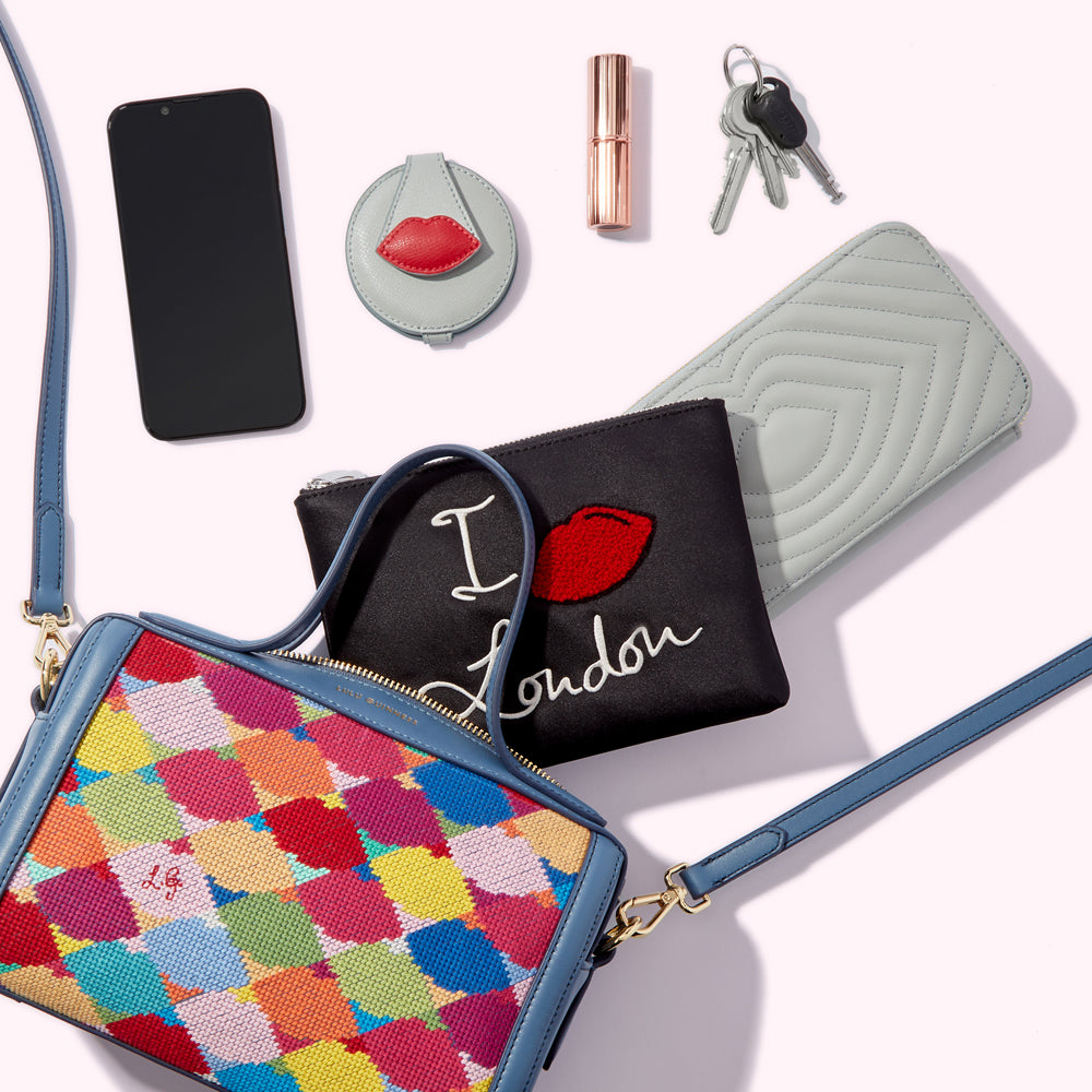 A colourful needlepoint handbag, a black "I love London" pouch, a light blue quilted wallet, a compact mirror with a red lip design, a lipstick, a smartphone, and keys are arranged on a white background.