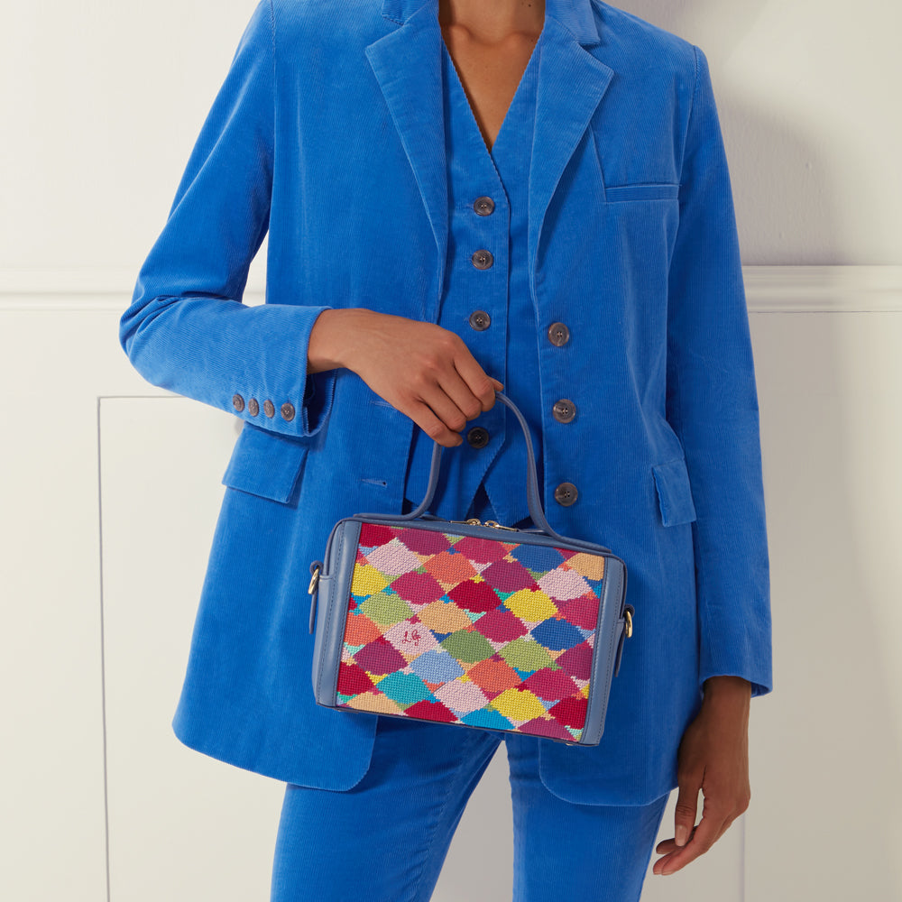 A woman in a blue corduroy suit holds a small, rectangular handbag with a multi-coloured, geometrically patterned front panel.