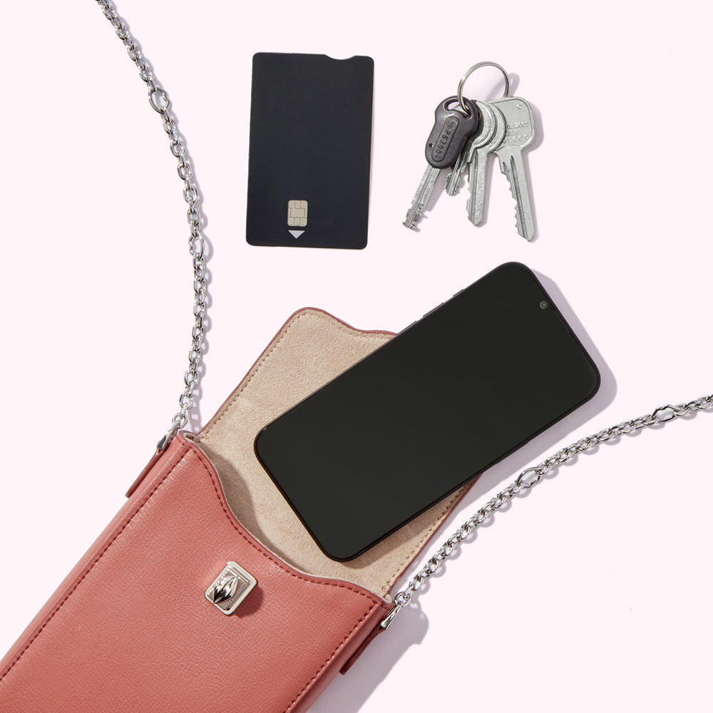 A blush pink leather crossbody phone bag with a silver chain strap, containing a smartphone and a beige microfiber interior. A black contactless payment card and a set of keys lie beside it on a white background.
