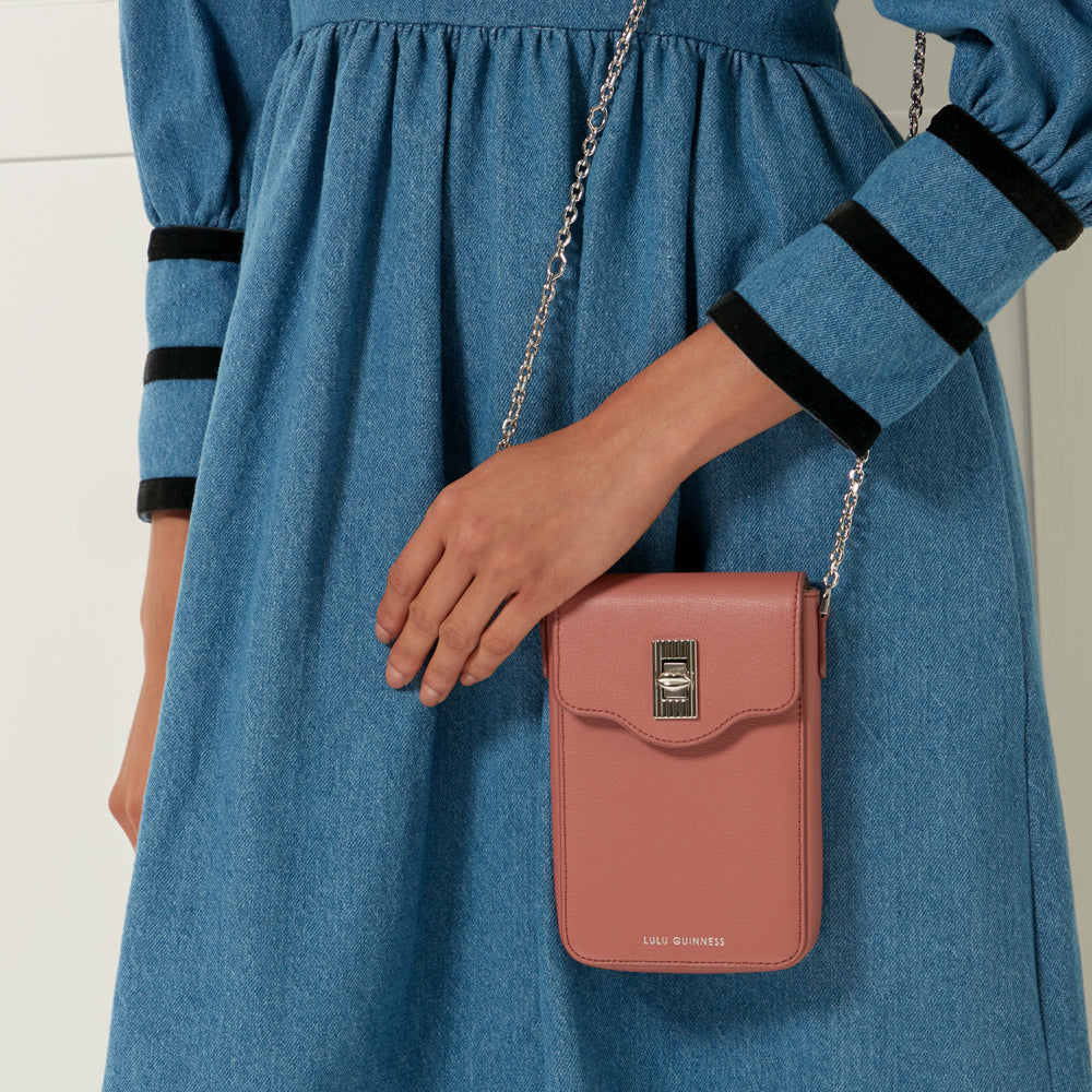 A dusty rose Lulu Guinness crossbody bag with a silver chain strap hangs from the shoulder of a person wearing a denim dress.