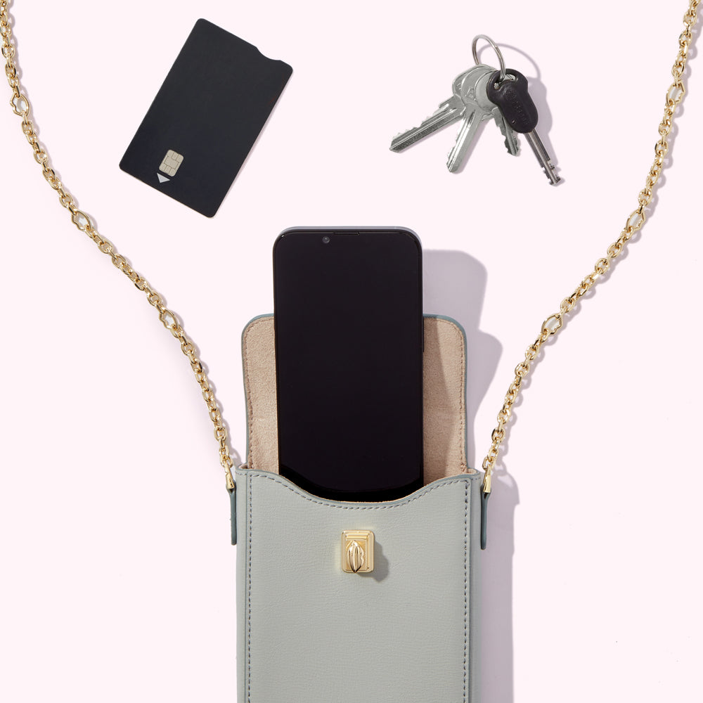 A light grey leather crossbody bag with a gold chain strap, containing a smartphone and a black card, sits on a white background, next to a set of keys.
