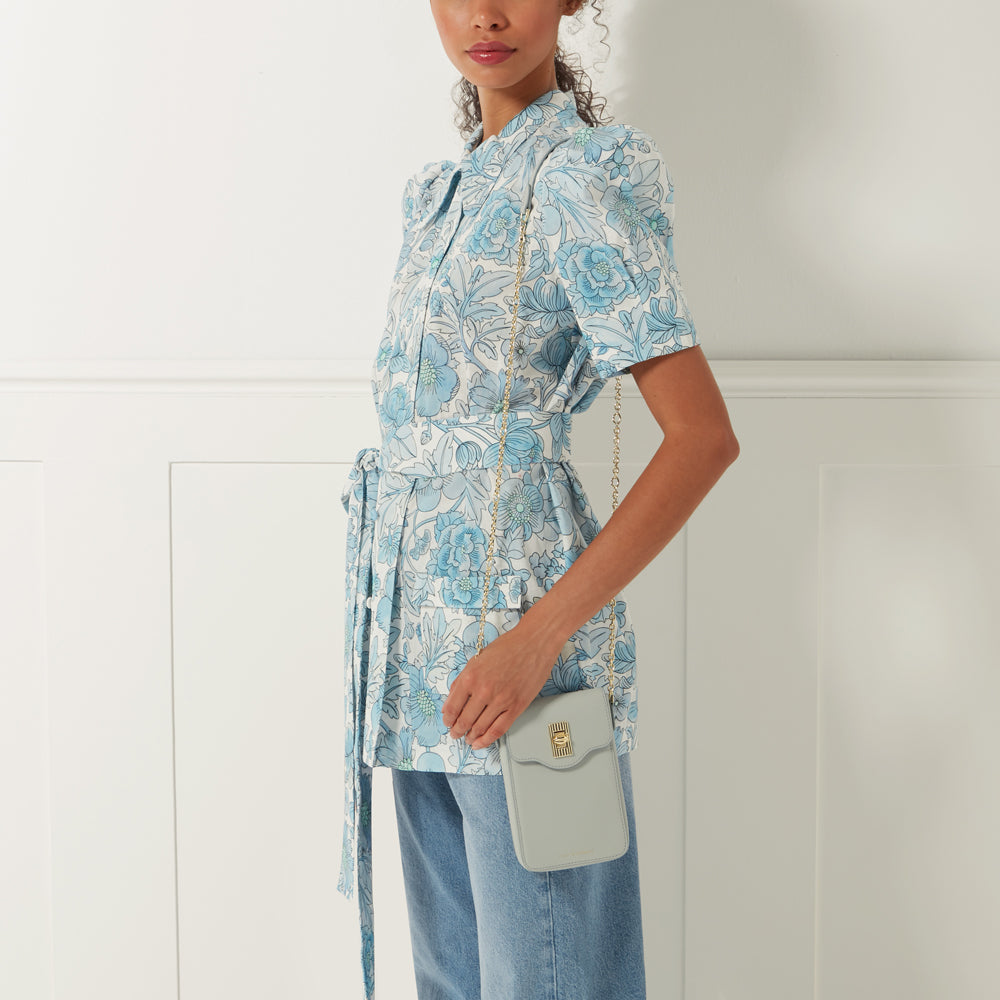 Woman in blue floral dress holding a light grey crossbody bag with gold chain strap against a white background.
