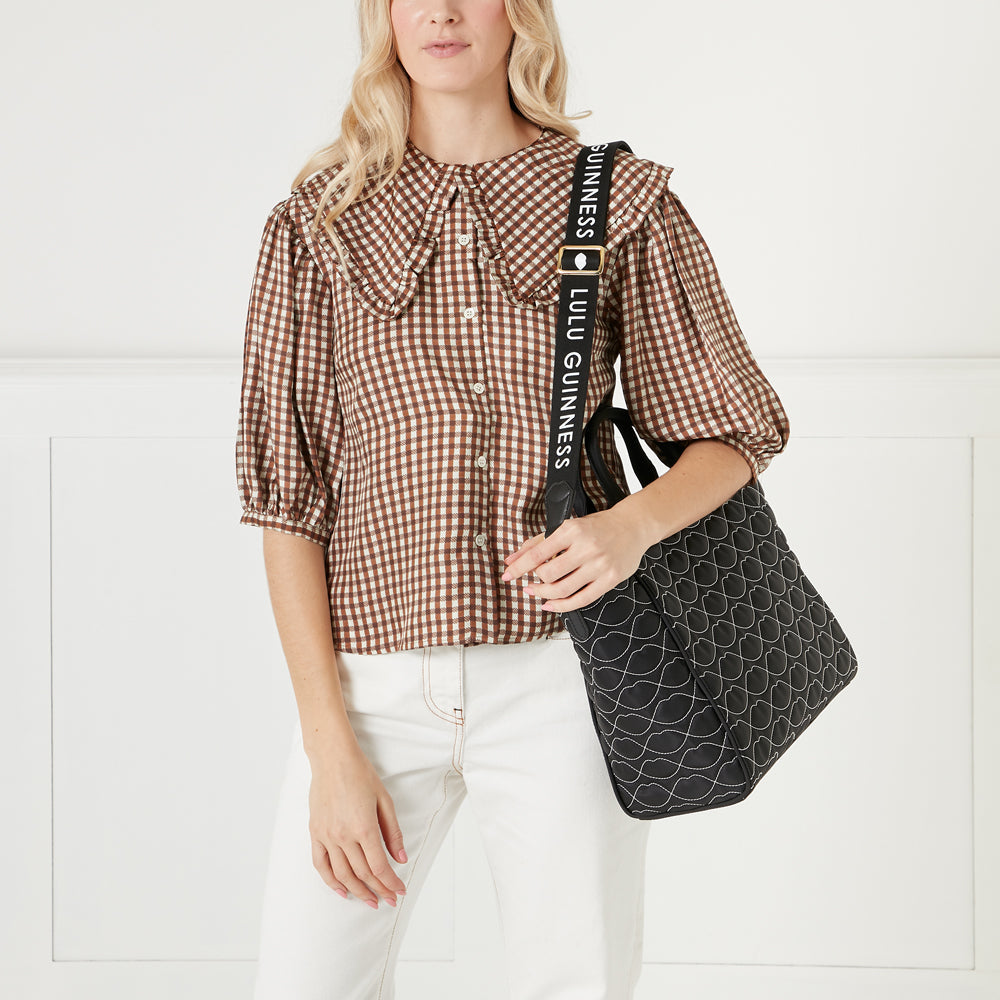 Woman in brown gingham shirt and white pants holding a black quilted Lulu Guinness tote bag with a black and white strap.
