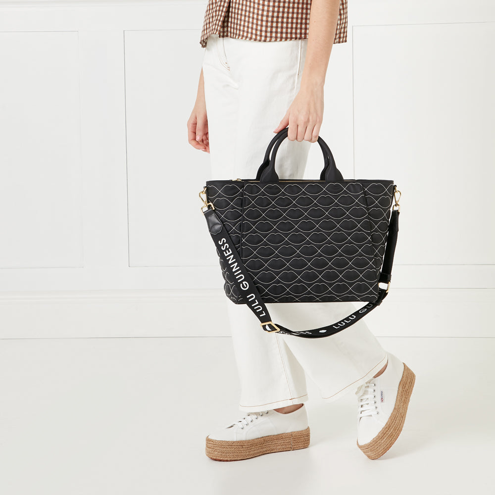 A woman in white pants holds a black Lulu Guinness tote bag with a lip print pattern and matching strap. The bag has gold hardware. She is wearing white platform espadrille sneakers. The background is white.
