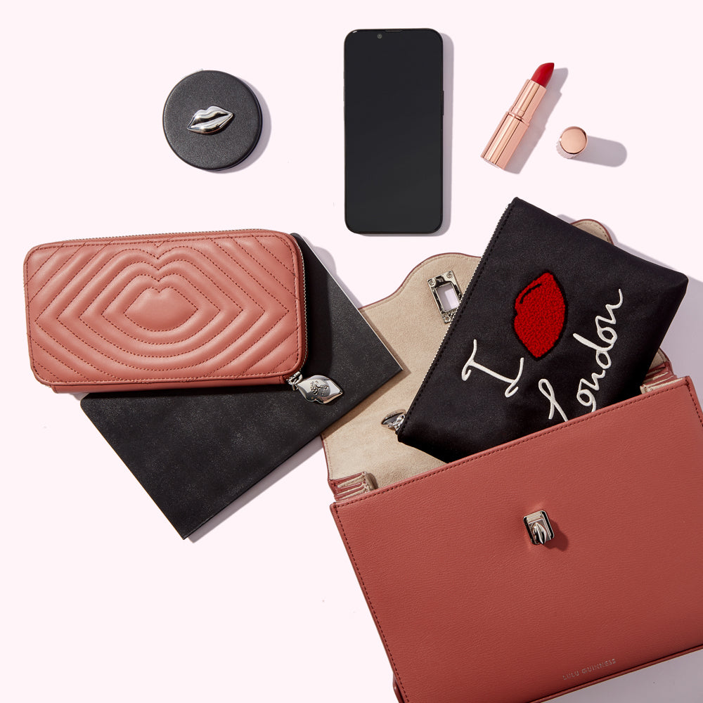 A blush pink handbag is open to reveal its contents: a pink quilted wallet, a black pouch with "I love London" embroidered on it, a black notebook, a smartphone, lipstick and a compact mirror. All items are arranged on a white background.
