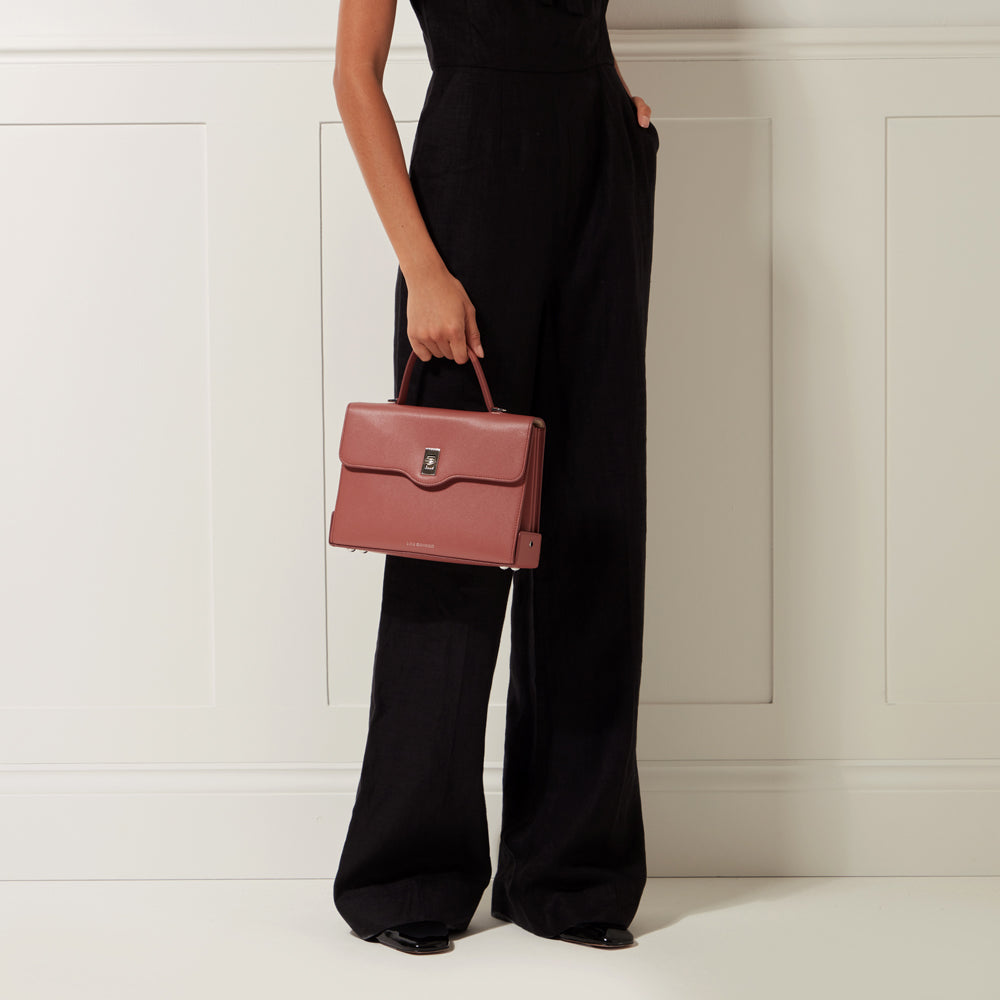 A woman in black wide-leg pants holds a dusty rose-coloured leather handbag against a white panelled background.
