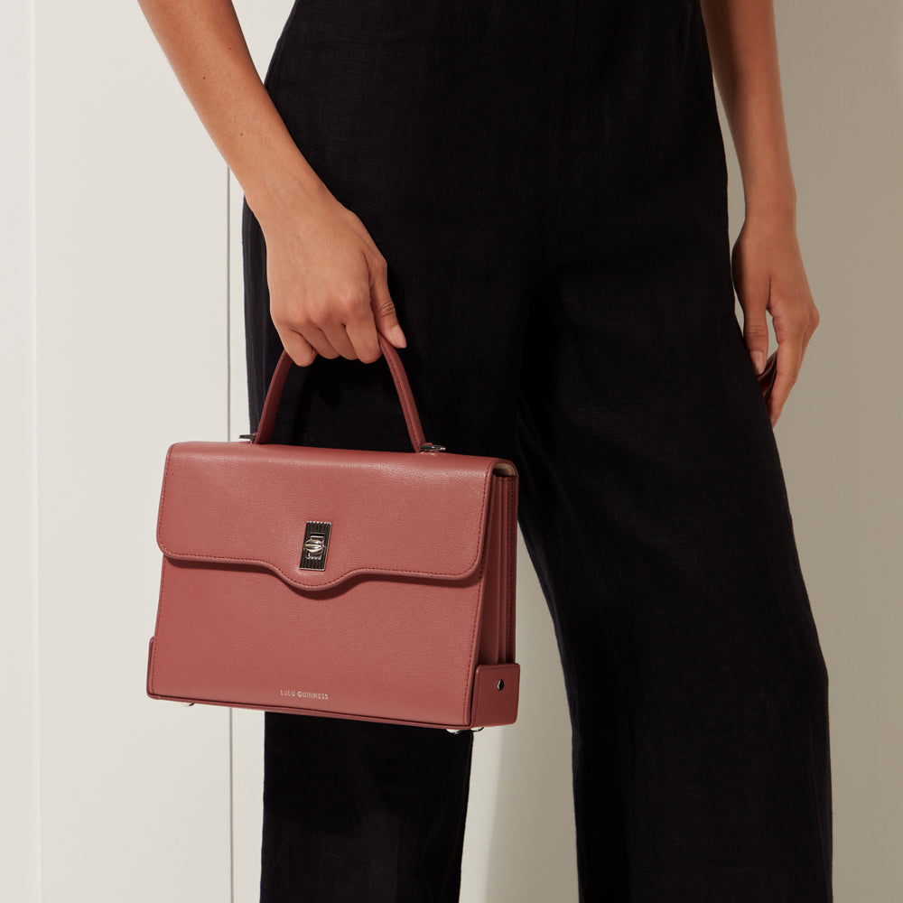 A dusty rose Lulu Guinness satchel held by a person wearing black pants against a white background.
