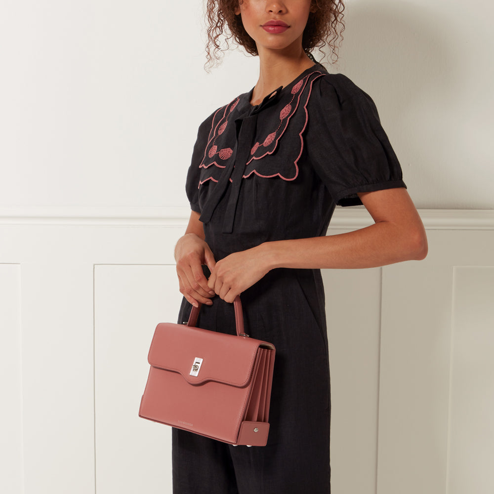 Woman in black dress with pink collar detail holding a dusty rose-coloured handbag against a white background.
