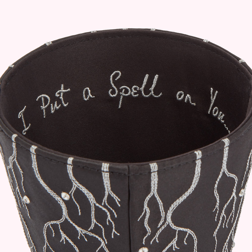 Close-up of a dark-coloured cylindrical container with silver embroidered roots on the exterior and the phrase "I Put a Spell on You" embroidered on the inside.
